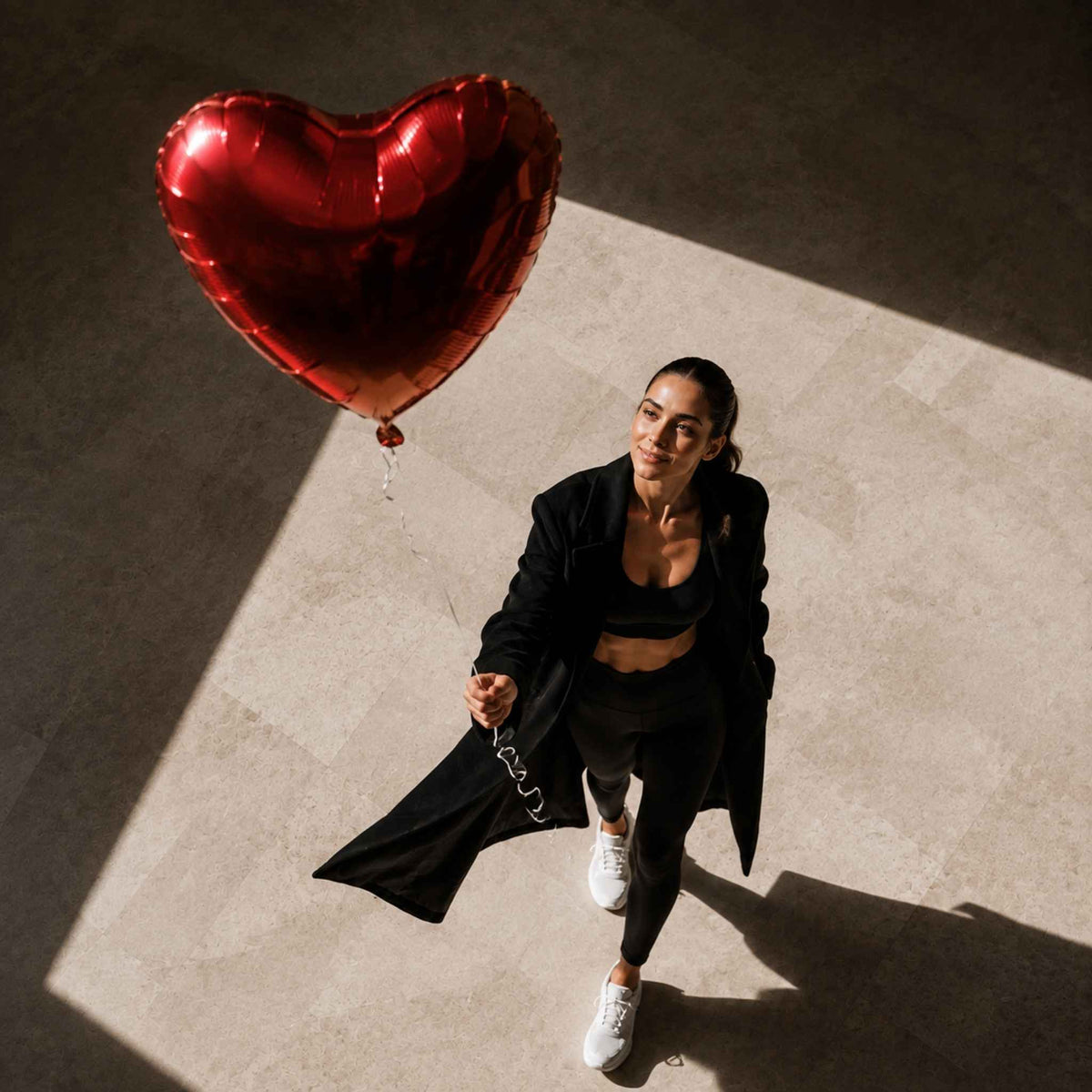 A woman in a black blazer, sports bra, and leggings walks across a sunlit concrete floor while holding a large metallic red heart-shaped balloon.
