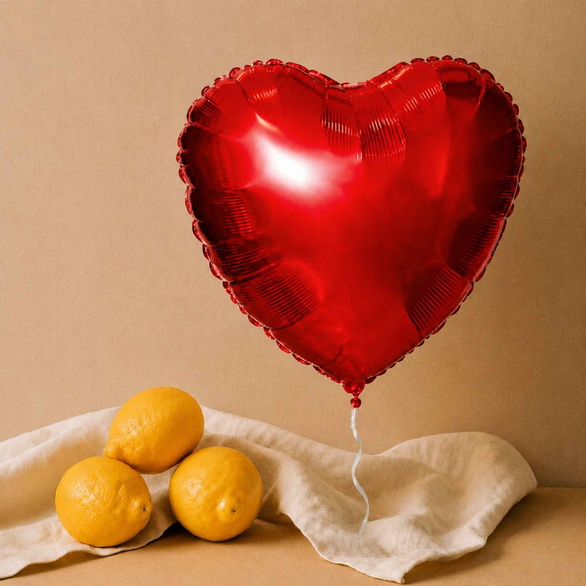 A shiny red heart-shaped foil balloon floating above three yellow lemons on a draped cream fabric against a warm beige background.