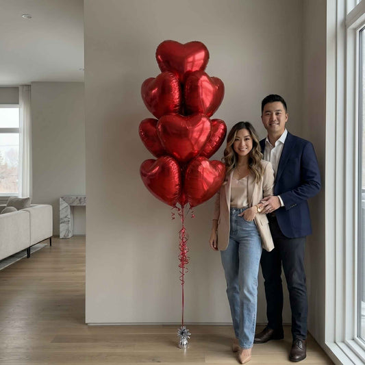 A husband and wife standing by the red foil balloon bouquet. Large apartment.