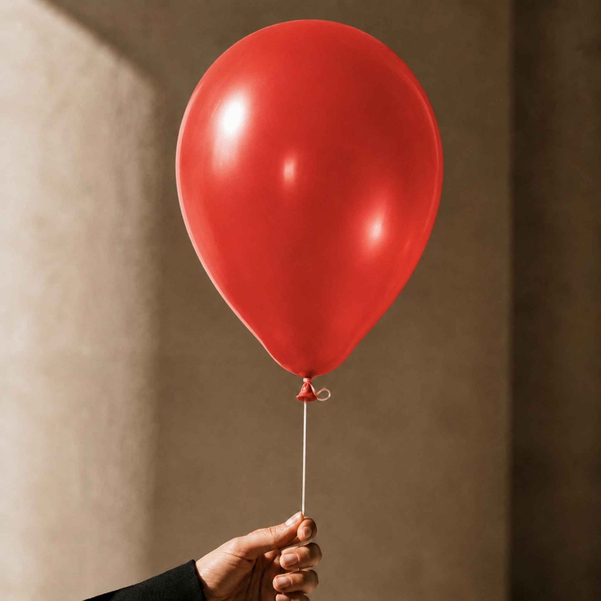 A single matte red balloon held by a hand against a warm beige wall with soft shadows.