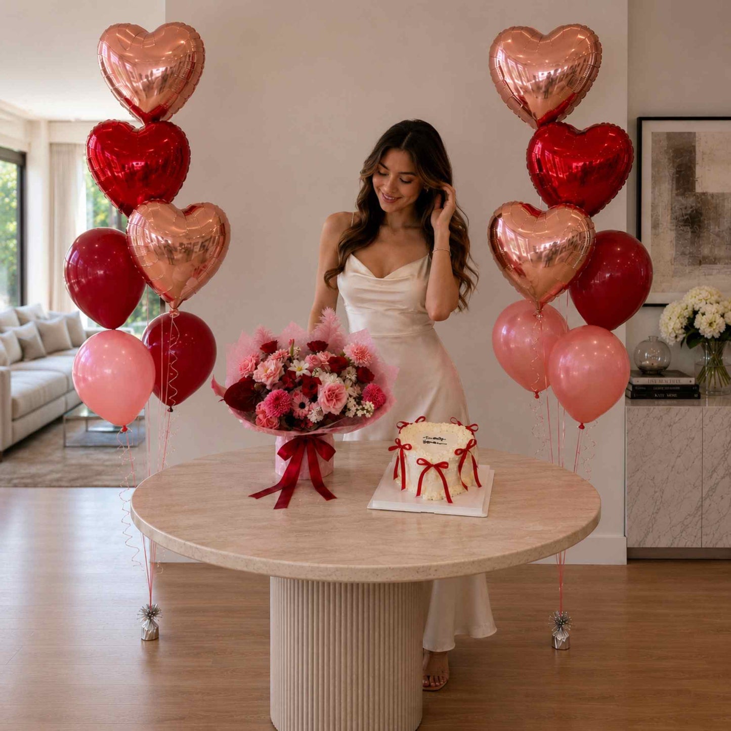 Woman in a white dress standing behind a table with balloons, flowers, and a cake in a living room.