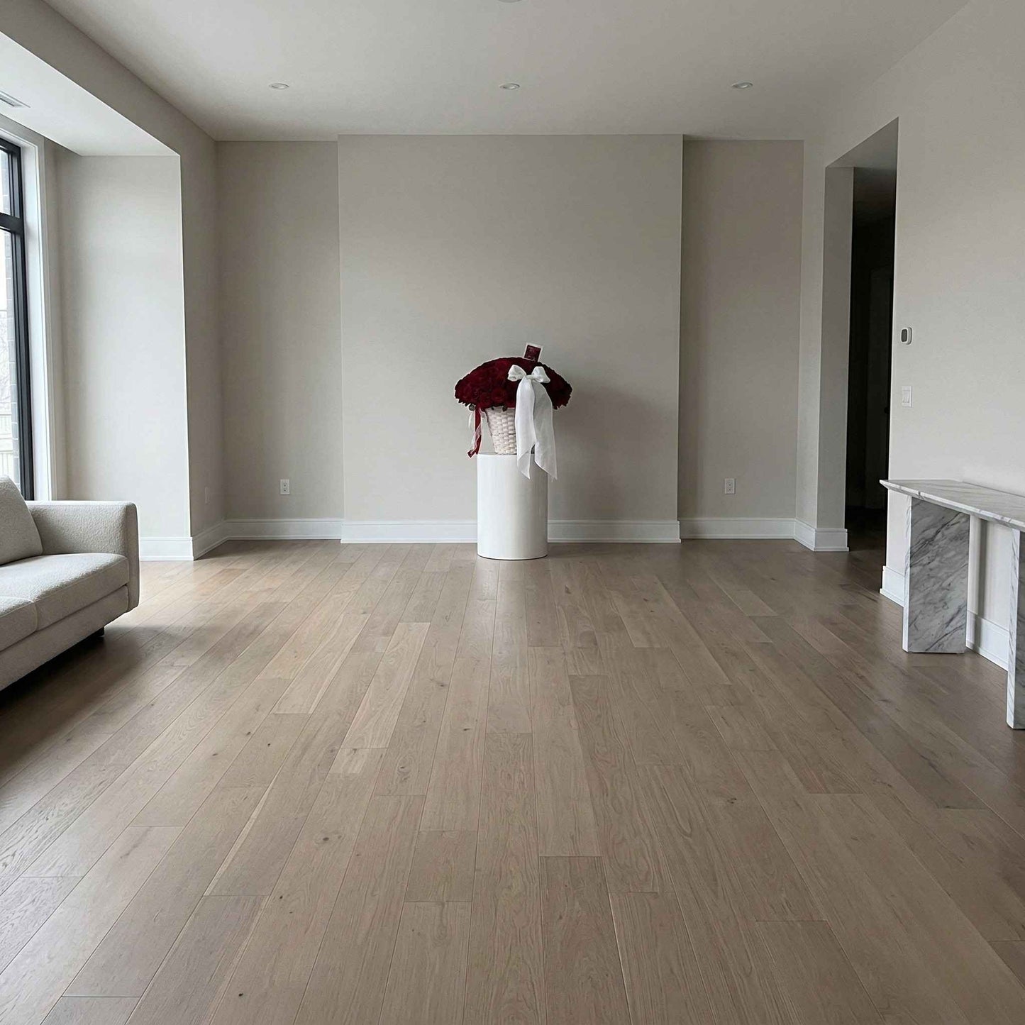 Empty living room with wooden flooring and a white vase with red and white flowers on a pedestal.