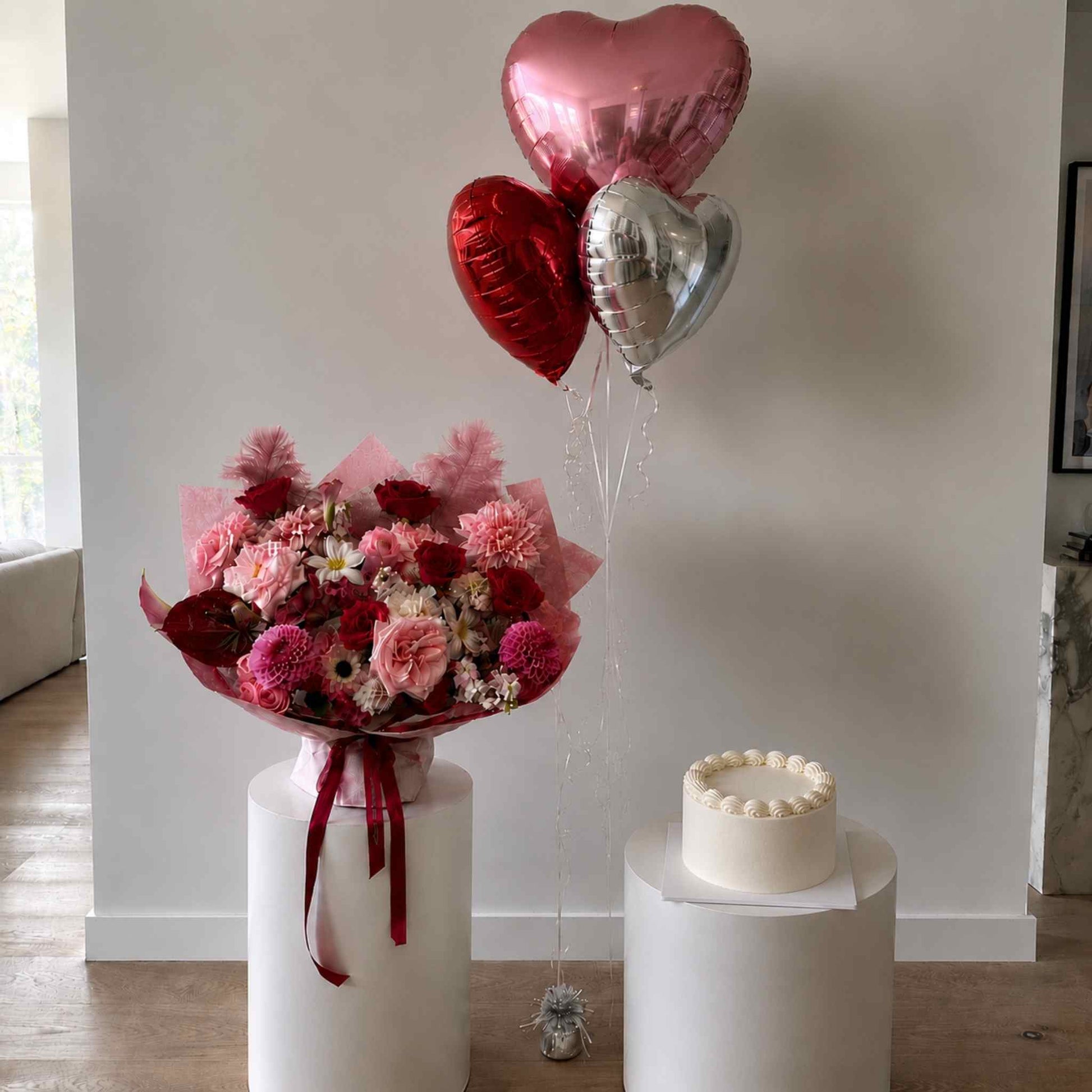 Bouquet of flowers with heart-shaped balloons and a small cake on white stands against a plain wall.