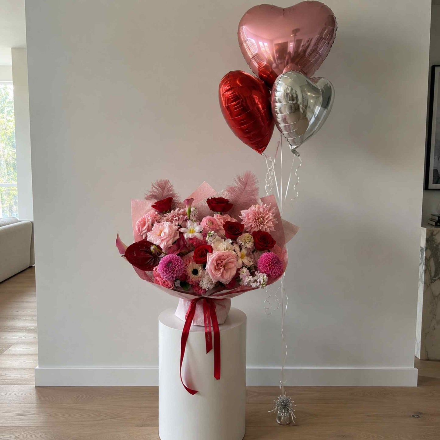 Bouquet of flowers with heart-shaped balloons on a white pedestal against a neutral wall.