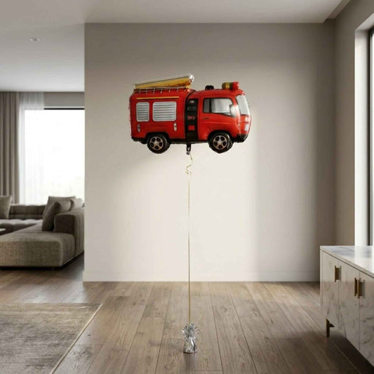 A photo taken in a modern, well-lit living room shows a red fire engine shaped foil helium balloon floating in the center. The balloon has black tires, silver details, and yellow and orange lights on its roof. A thin gold ribbon extends straight down from the balloon to a shiny silver balloon weight on the floor.