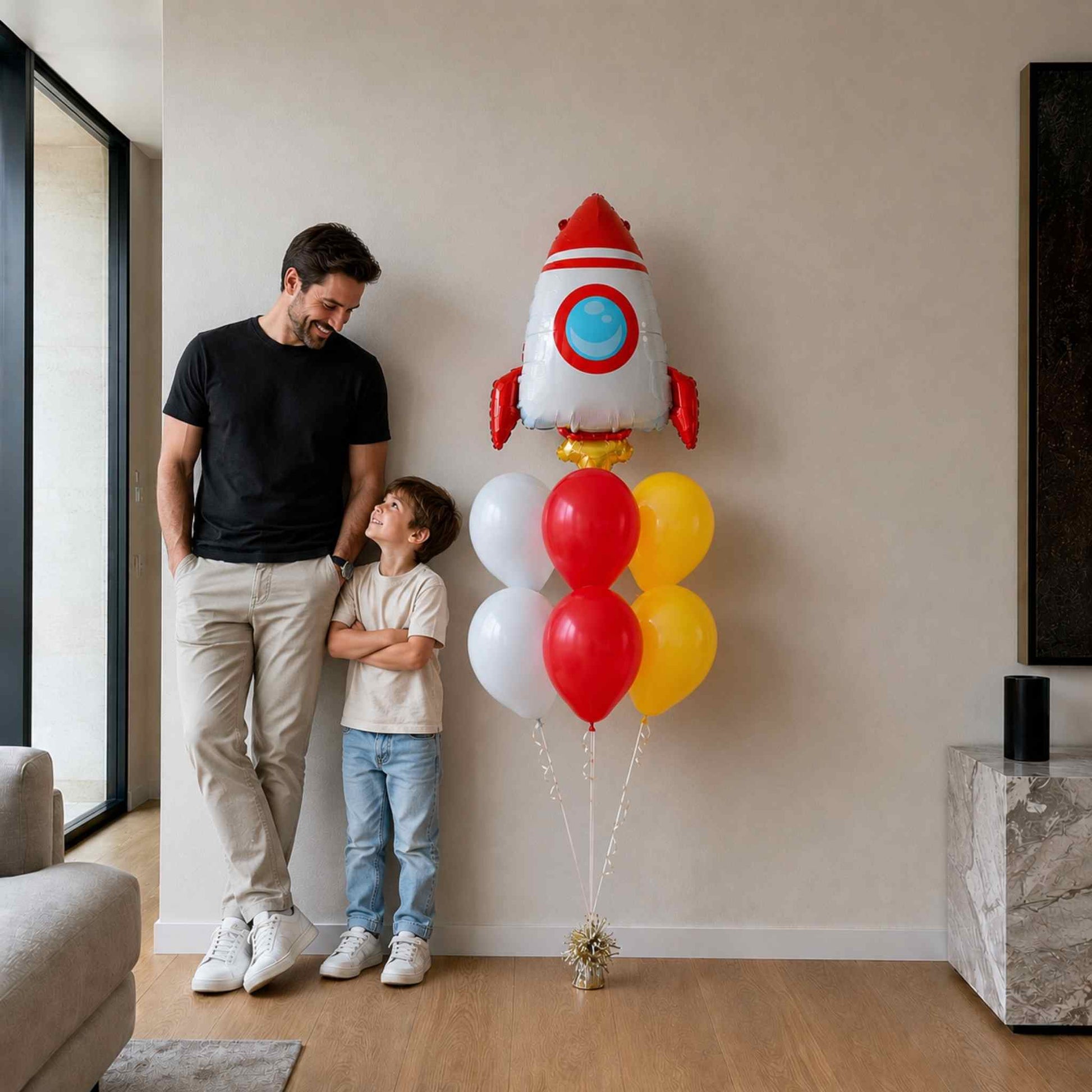 Man and child standing next to a rocket-shaped balloon in a living room.