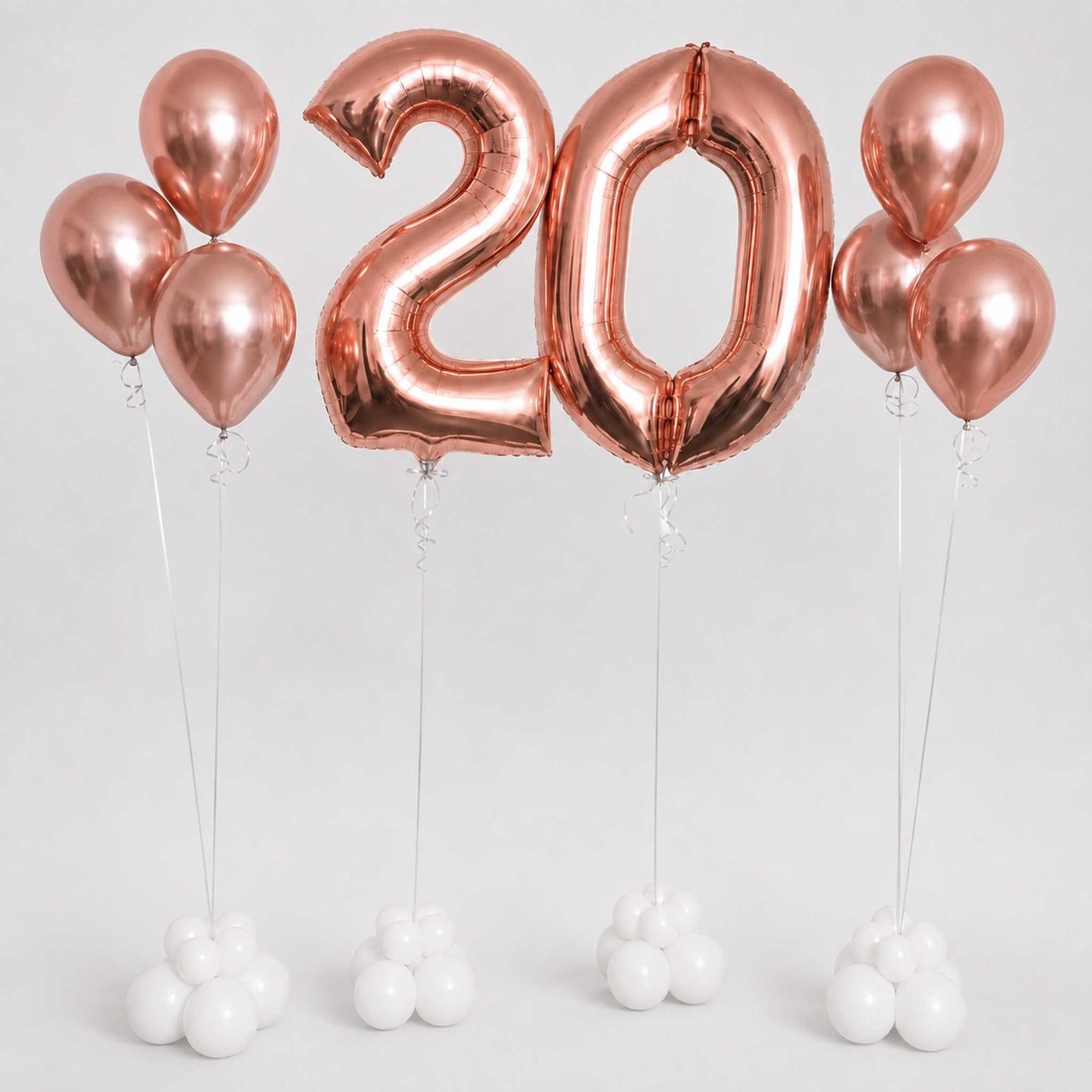 Rose gold foil balloons spelling out '20' with matching round balloons on strings, arranged on a light gray background with small white balloons at the base.