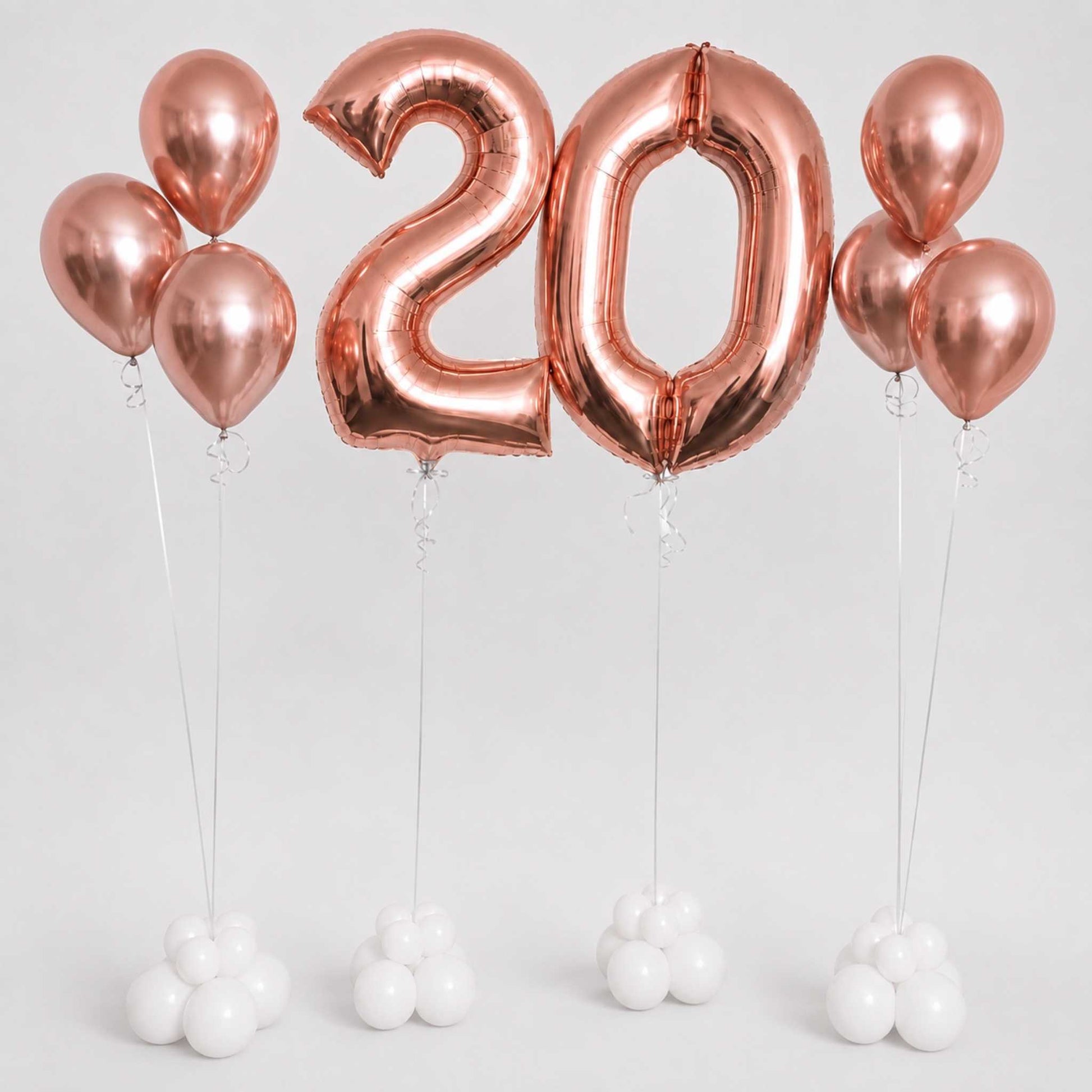 Rose gold foil balloons spelling out '20' with matching round balloons on strings, arranged on a light gray background with small white balloons at the base.