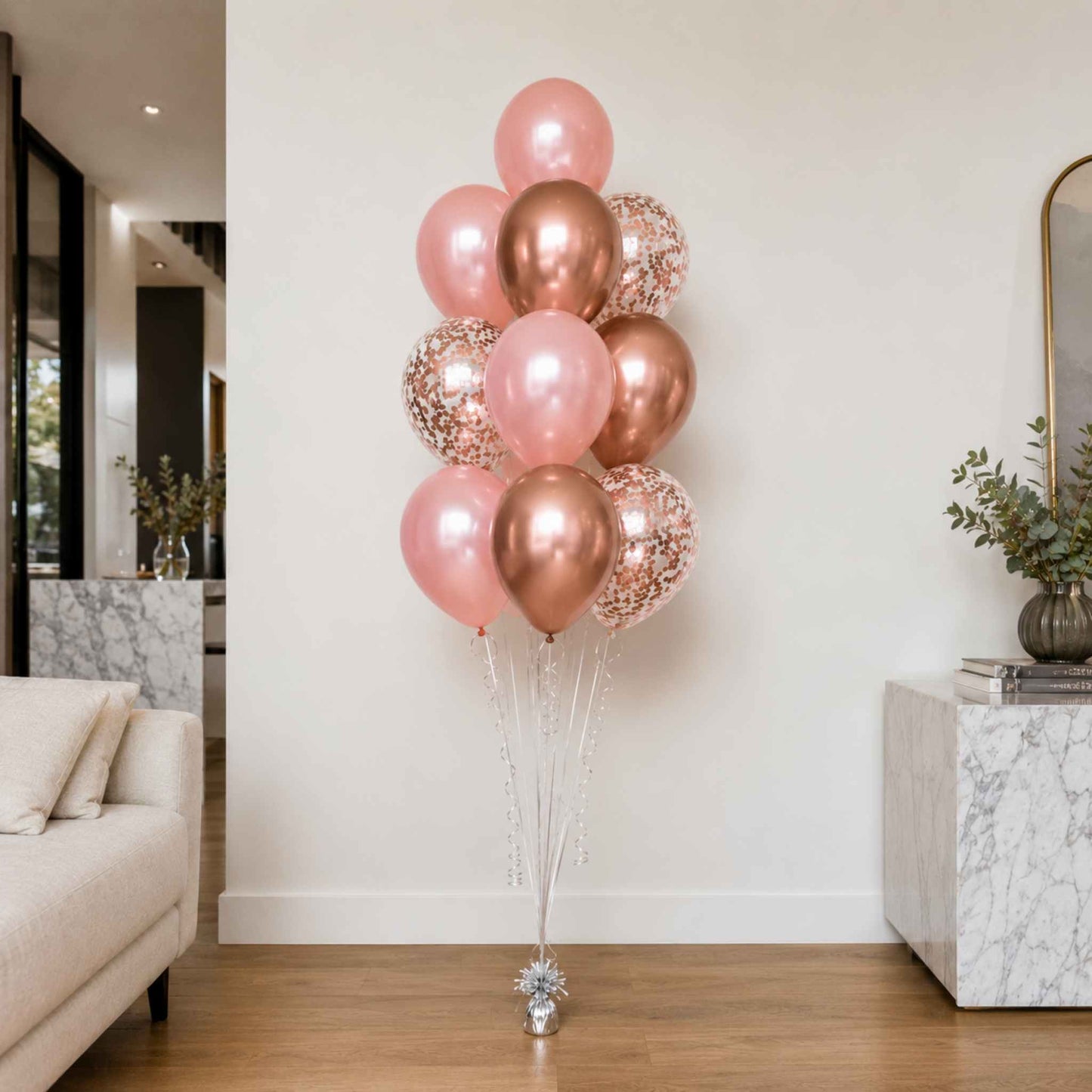 A cluster of rose gold and pink balloons, including metallic, foil, and confetti designs, tied to a silver stand in a modern living room setting.
