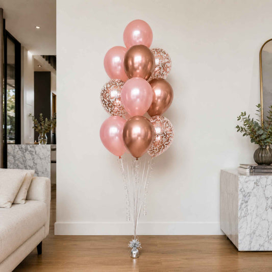 A cluster of rose gold and pink balloons, including metallic, foil, and confetti designs, tied to a silver stand in a modern living room setting.