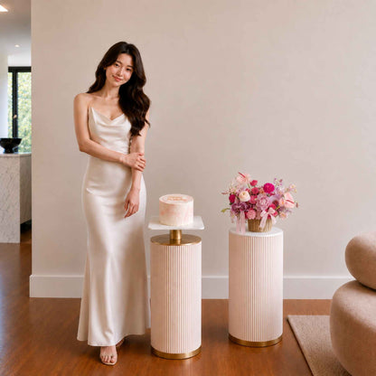 Woman in a white dress standing next to a small cake and floral arrangement on a wooden floor.