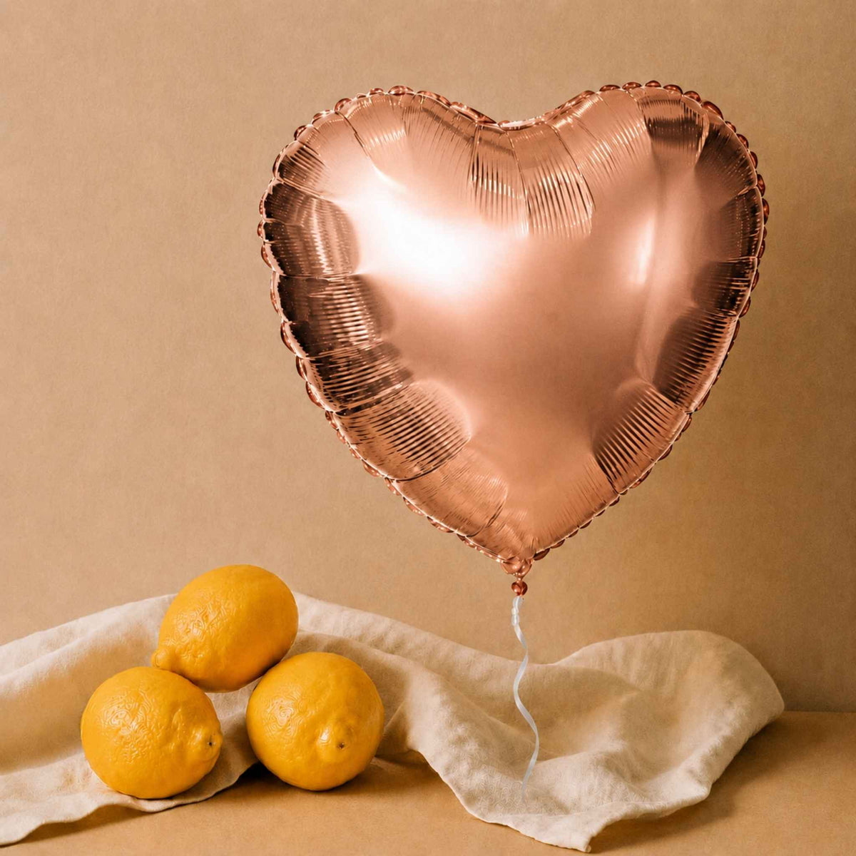 A metallic rose gold heart-shaped foil balloon floating above three yellow lemons on a draped cream linen cloth against a warm beige background.