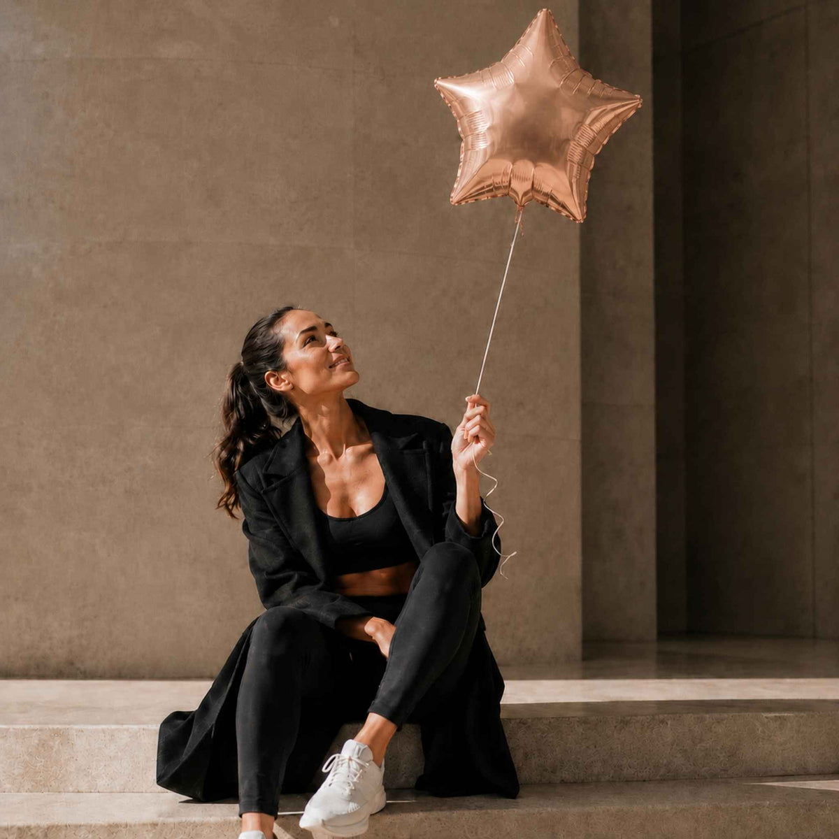A woman in a black blazer, sports bra, and leggings sits on stone steps holding a metallic rose gold star-shaped foil balloon against a textured concrete wall.