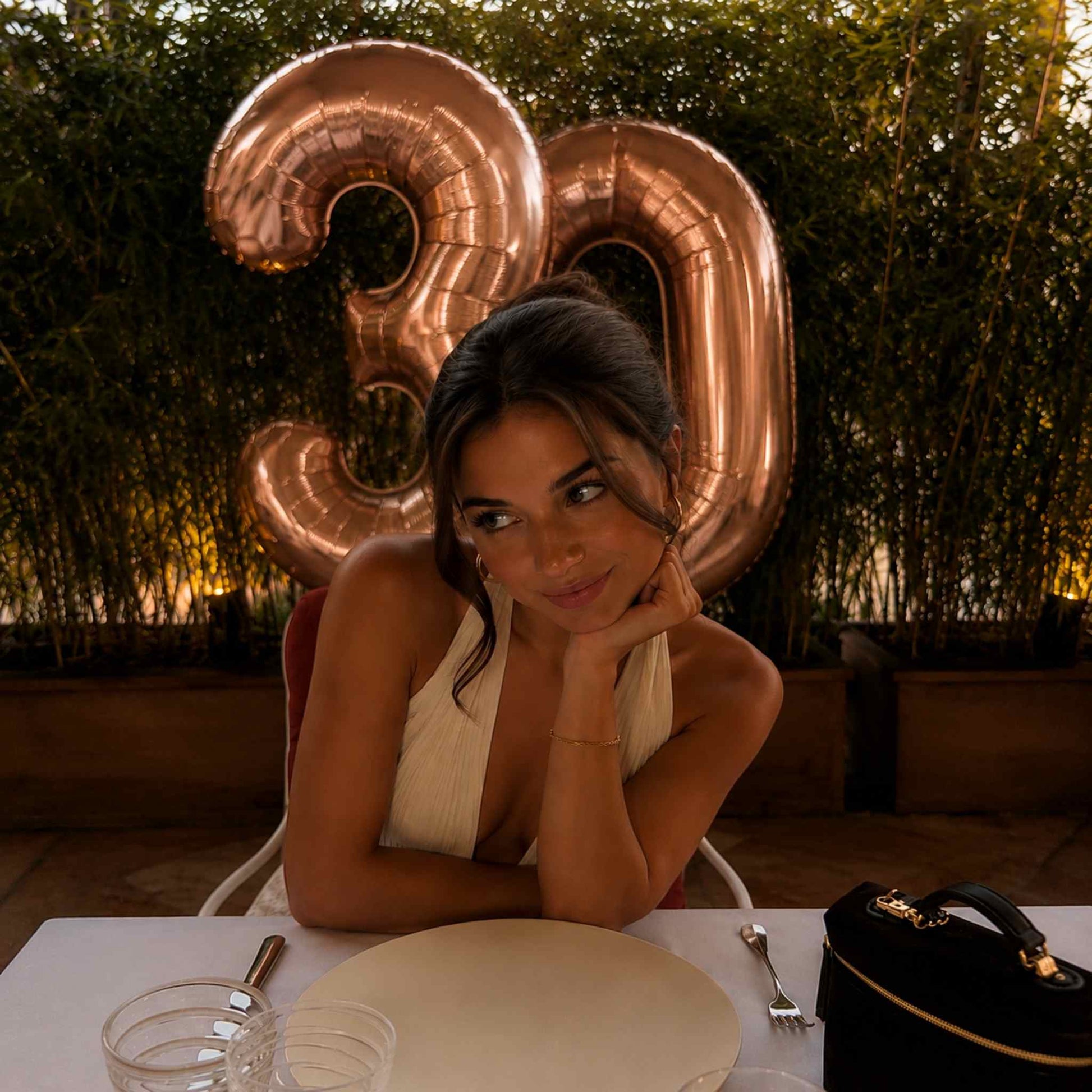 A woman in a cream halter dress sits at a table with a large metallic rose gold balloon number 30 behind her, with a black handbag in the foreground.