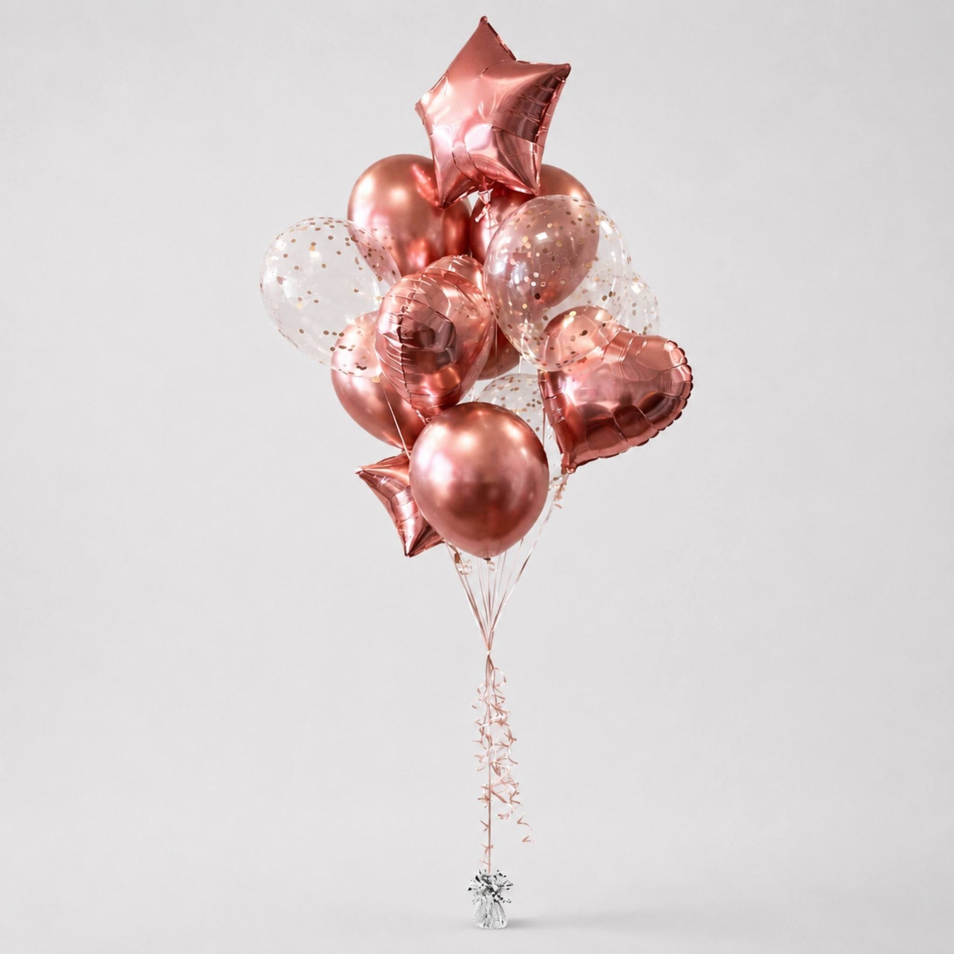 A cluster of metallic rose gold foil balloons in star, heart, and round shapes, mixed with clear balloons containing gold confetti, tied together with a decorative string and a small silver star finial, on a light gray background.
