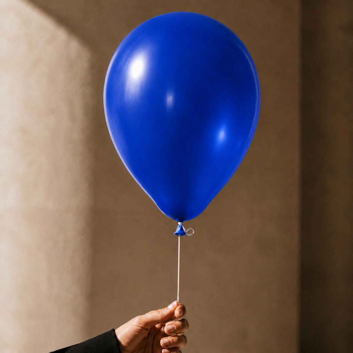 A single glossy royal blue balloon held by a hand against a warm beige wall.