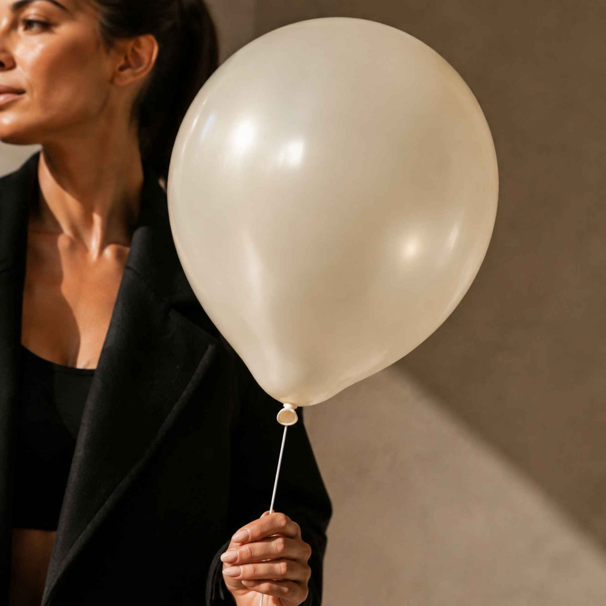 A woman in a black blazer holds a large, unbleached off-white latex balloon against a warm, neutral-toned background.