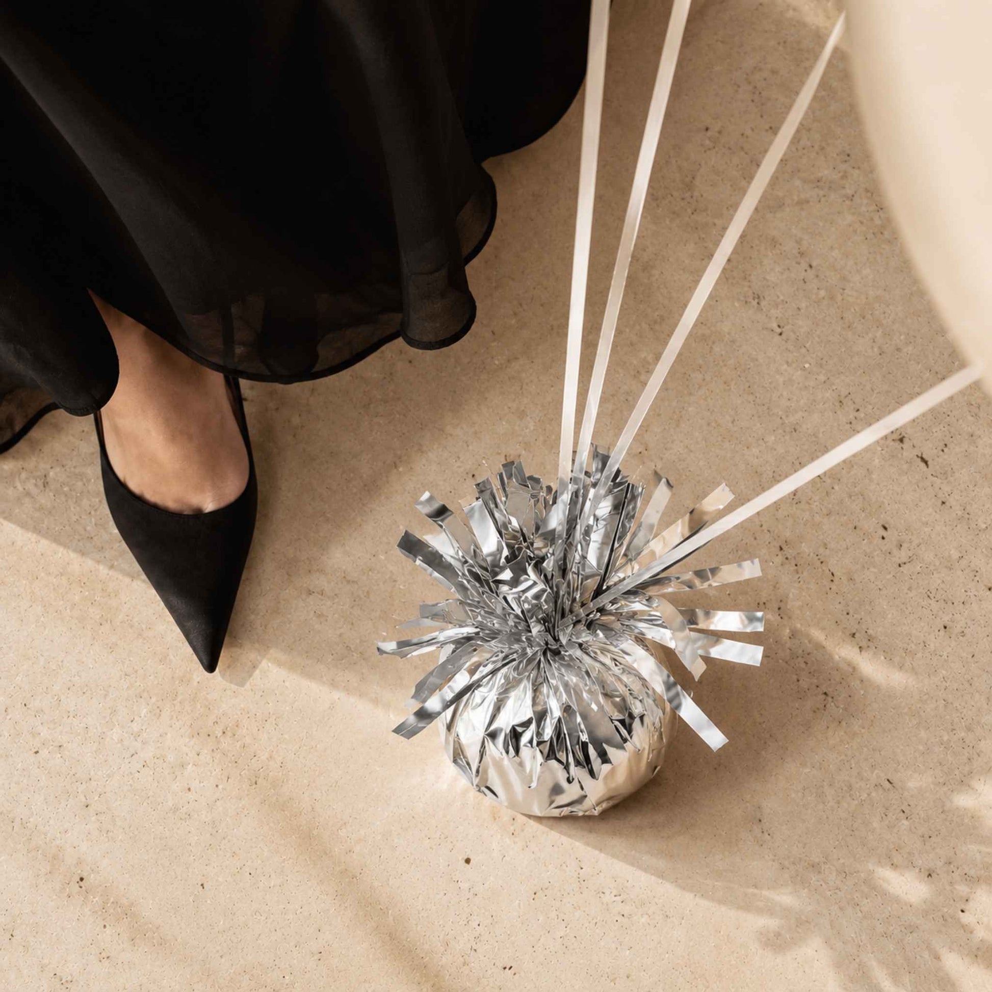 A silver metallic tinsel ball decoration with white ribbons, placed on a beige stone floor next to a person's feet wearing black pointed flats and a black skirt.