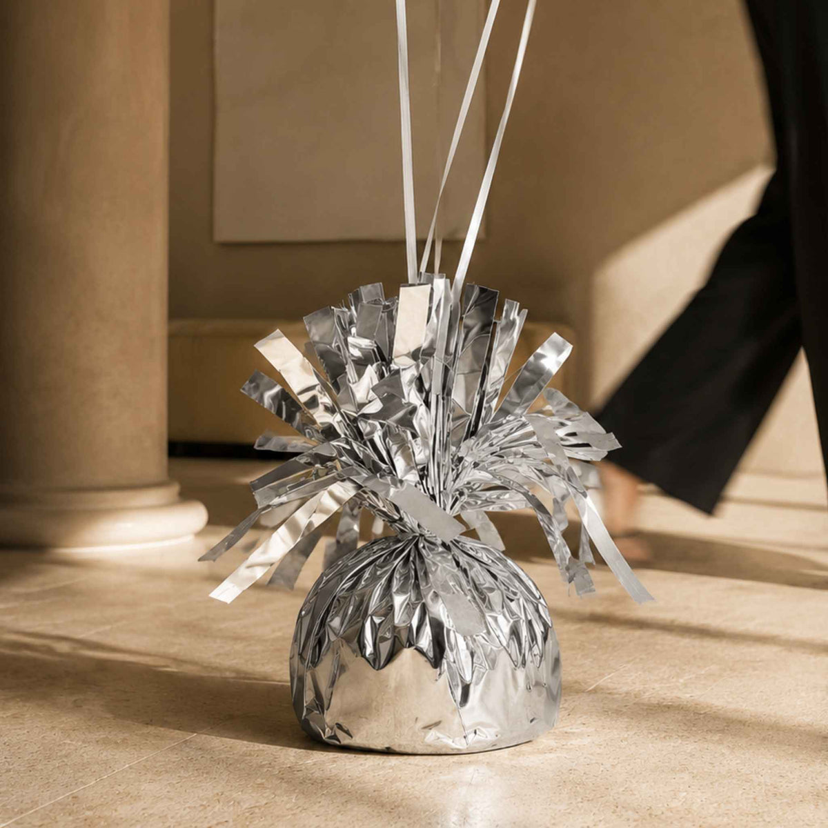 A silver metallic tinsel burst decoration with white hanging ribbons, placed on a beige stone floor in an indoor setting with a blurred person and column in the background.