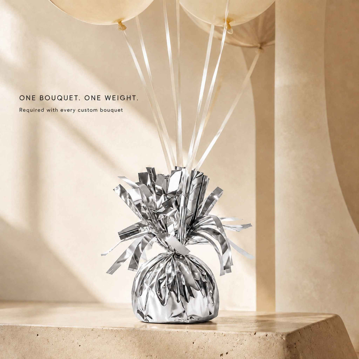 A silver foil-wrapped bouquet with metallic tinsel ribbon and long white string balloons, displayed on a beige stone surface against a warm beige architectural background. Text on the left reads, 'ONE BOUQUET.