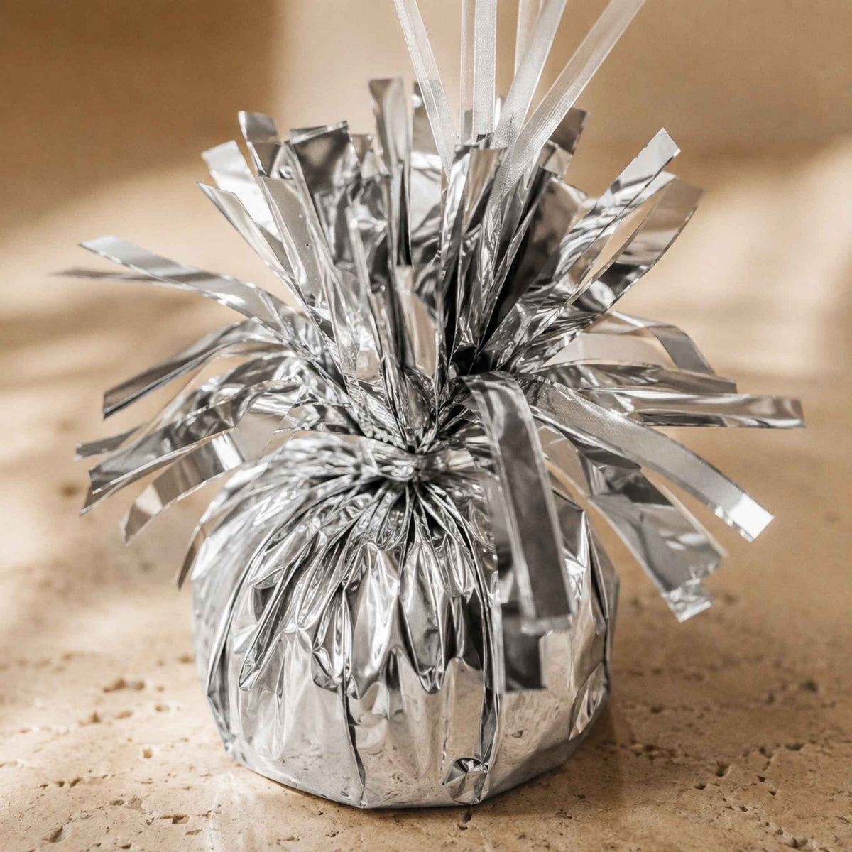 A small silver metallic tinsel decoration with a rounded base and radiating foil strands, photographed on a warm beige textured surface.
