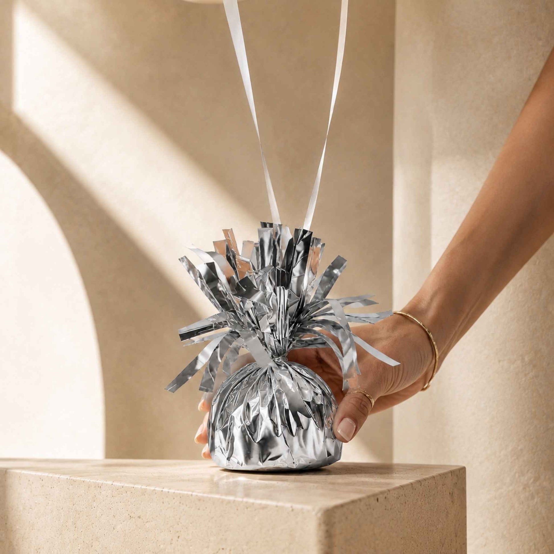 A hand holding a shiny silver foil party decoration shaped like a small bag with metallic tinsel streamers and white ribbon strings, set against a warm beige architectural background.