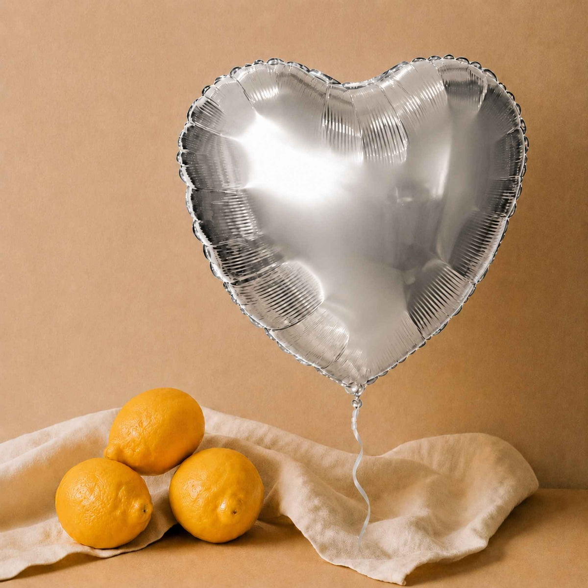 A metallic silver heart-shaped foil balloon floating above three yellow lemons on a beige cloth against a warm tan background.