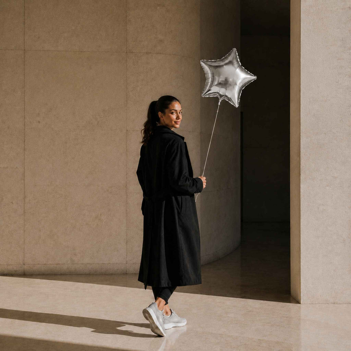 A woman in a long black coat and white sneakers stands in a sunlit stone corridor, holding a metallic silver star-shaped balloon.