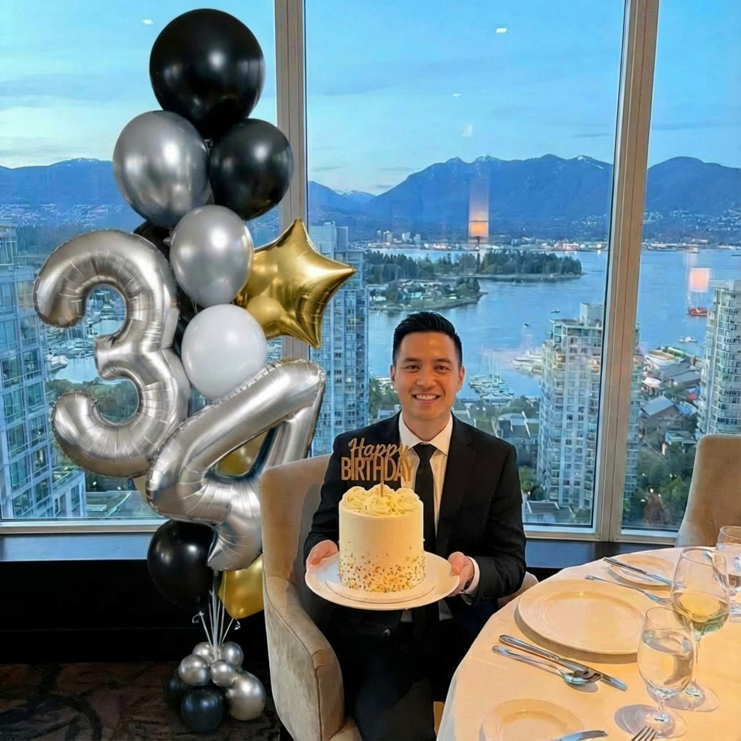 Man holding a birthday cake with balloons and a scenic city view in the background