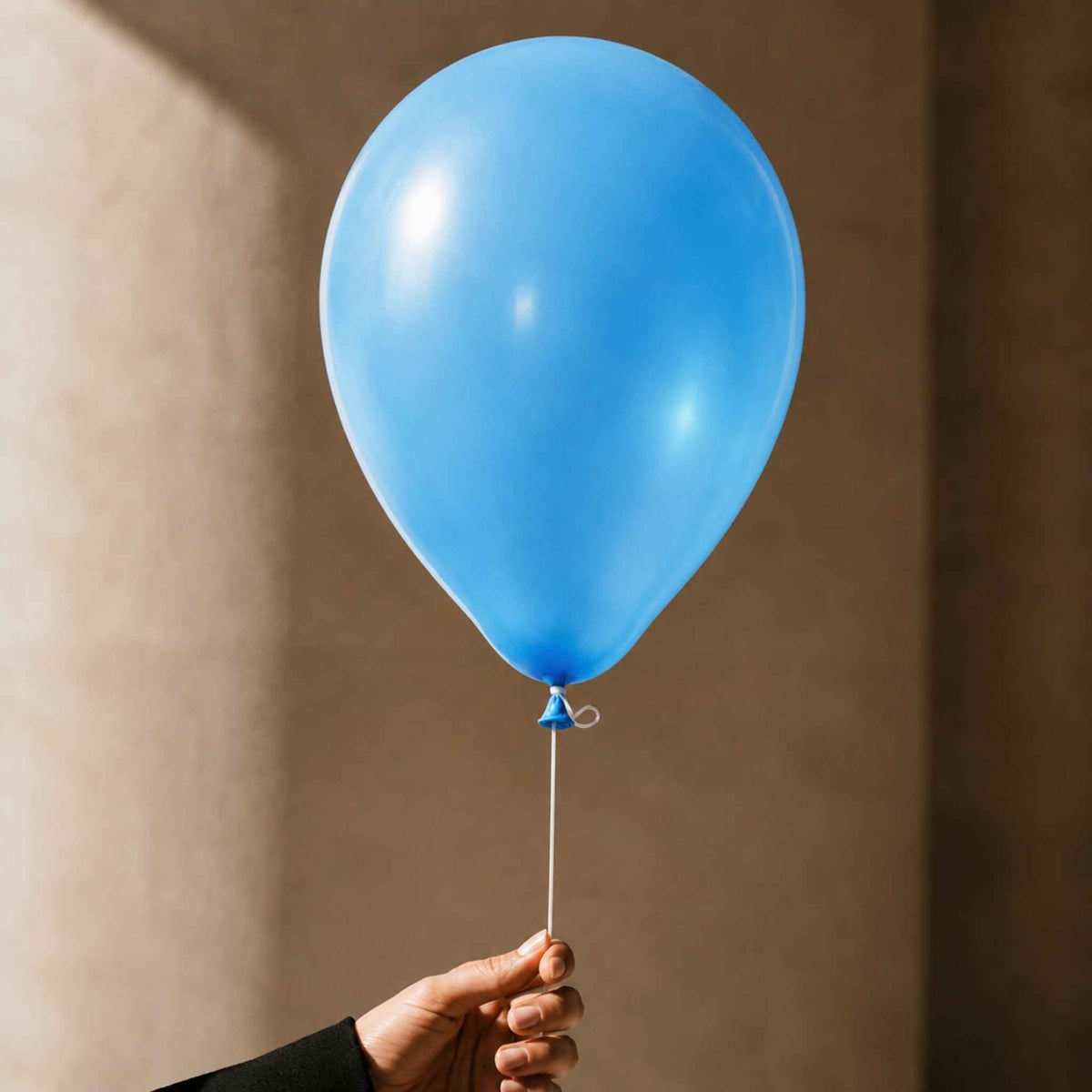 A single light blue latex balloon held by a hand against a warm beige wall background.
