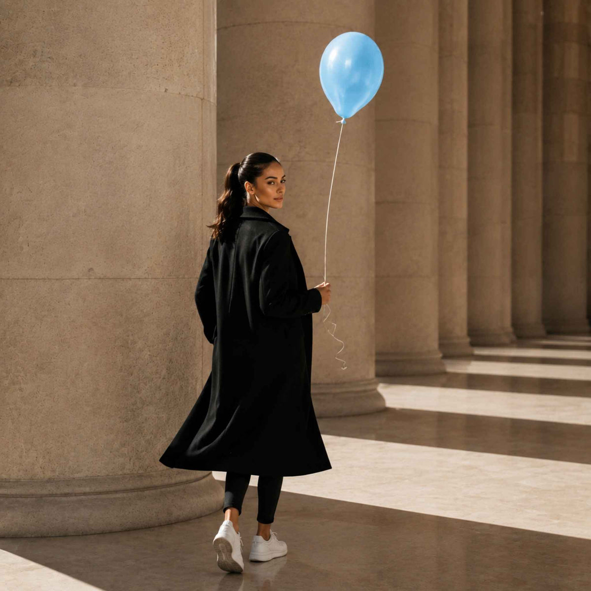 A woman in a long black coat stands in a grand columned hall, holding a light blue balloon. She is wearing black leggings and white sneakers.