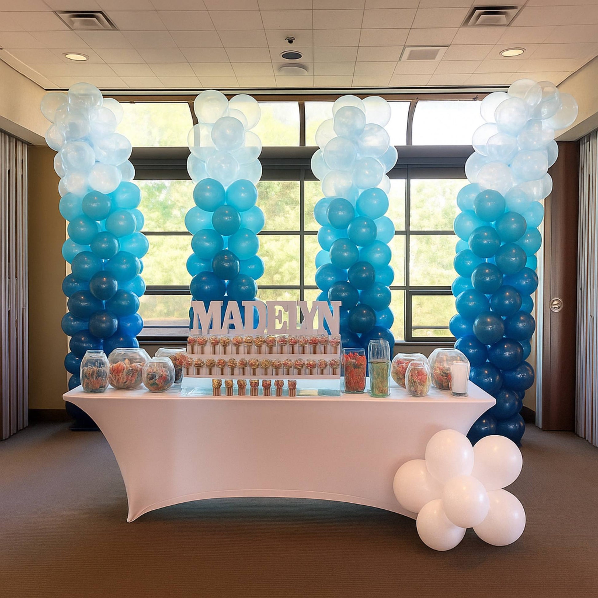 Decorative balloon arch with 'MADELYN' sign above a dessert table in a room with large windows.