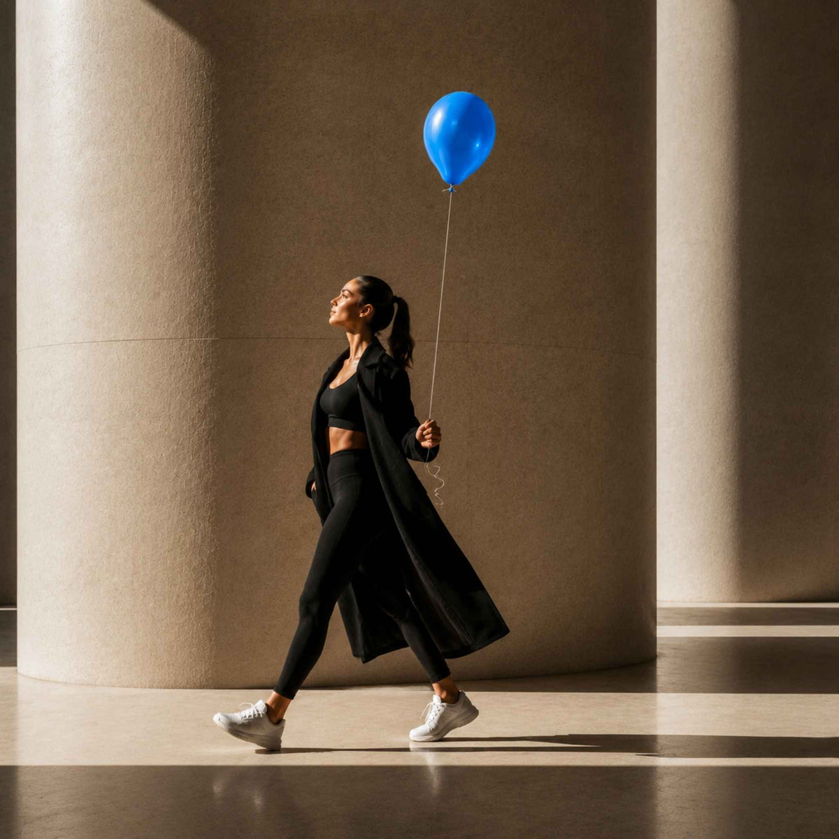 A woman in a black activewear set with a long black coat walks past large beige columns while holding a bright blue balloon.