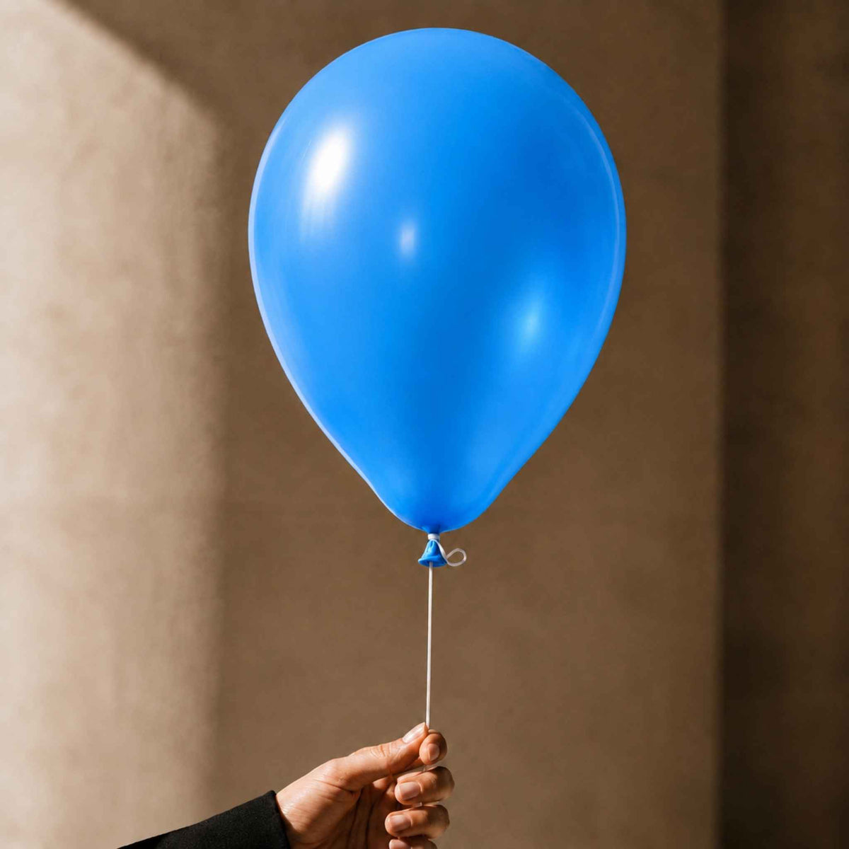 A single bright blue latex balloon held by a hand against a warm beige wall background.