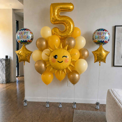 Balloon arrangement with a number 5 gold balloon, sun-shaped balloon, and 'Happy Birthday' balloons in a room.