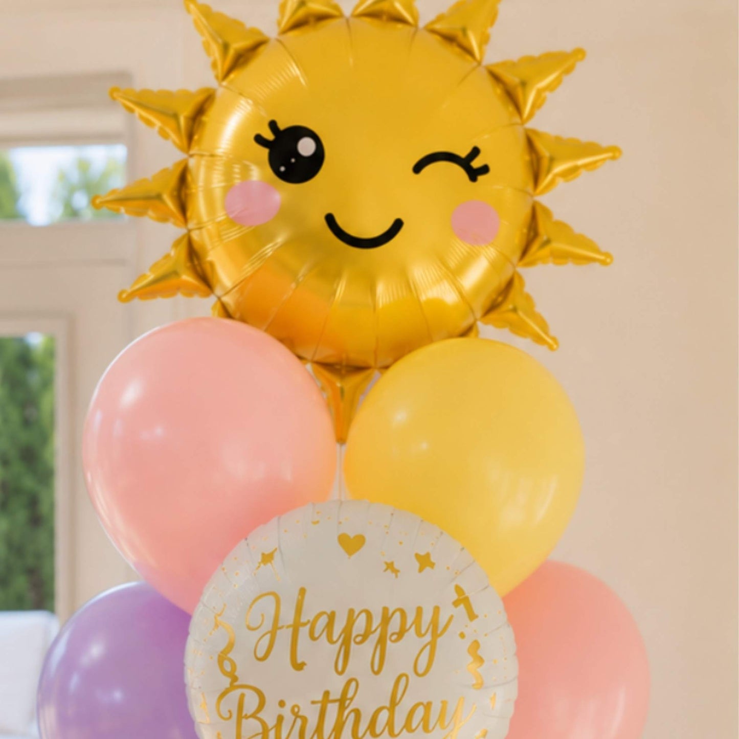 A birthday balloon bouquet featuring a large metallic gold foil sun balloon with a winking face, pink and yellow latex balloons, and a round white foil balloon printed with 'Happy Birthday' in gold script.