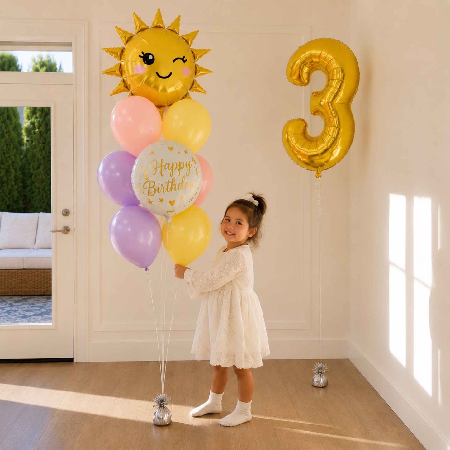 A young girl in a white dress stands indoors holding a balloon bouquet with a large gold sun balloon, pastel pink, yellow, and lavender balloons, and a gold number 3 balloon on a string. The room has cream walls, wood floors, and a glass door in the background.