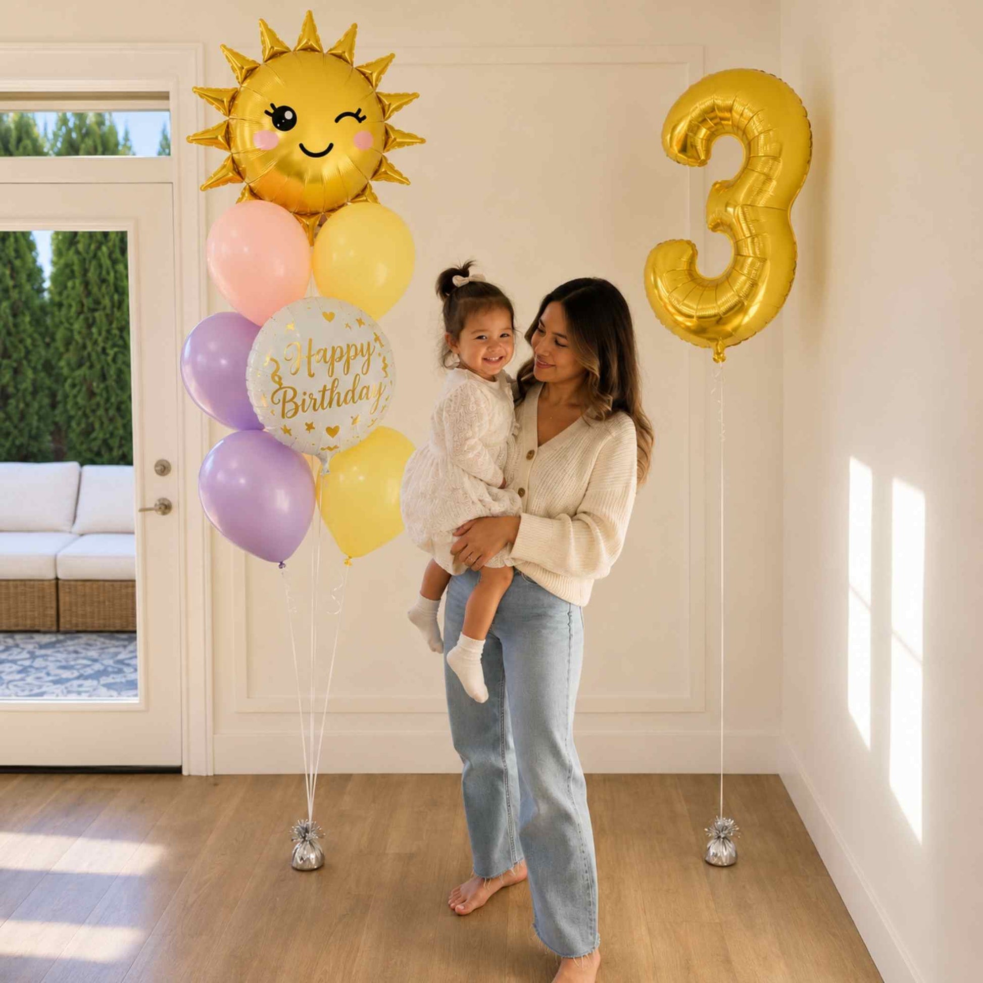 A woman holding a young child in a bright room decorated for a birthday, with a gold foil balloon shaped like the number 3, a cluster of pastel balloons including lavender, pink, yellow, and a white balloon that says 'Happy Birthday,' and a large winking sun balloon.