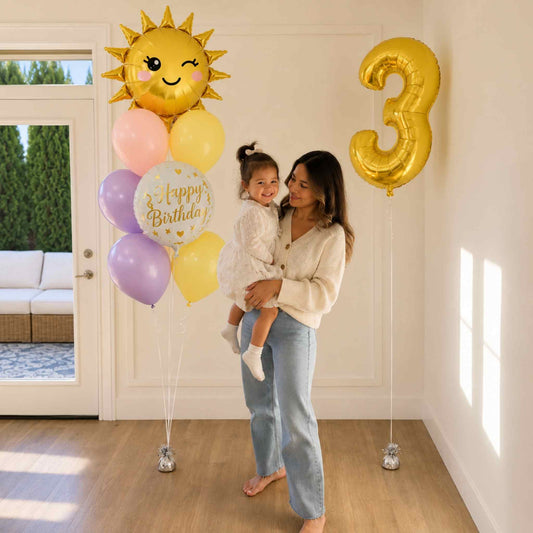A woman holding a young child in a bright room decorated for a birthday, with a gold foil balloon shaped like the number 3, a cluster of pastel balloons including lavender, pink, yellow, and a white balloon that says 'Happy Birthday,' and a large winking sun balloon.