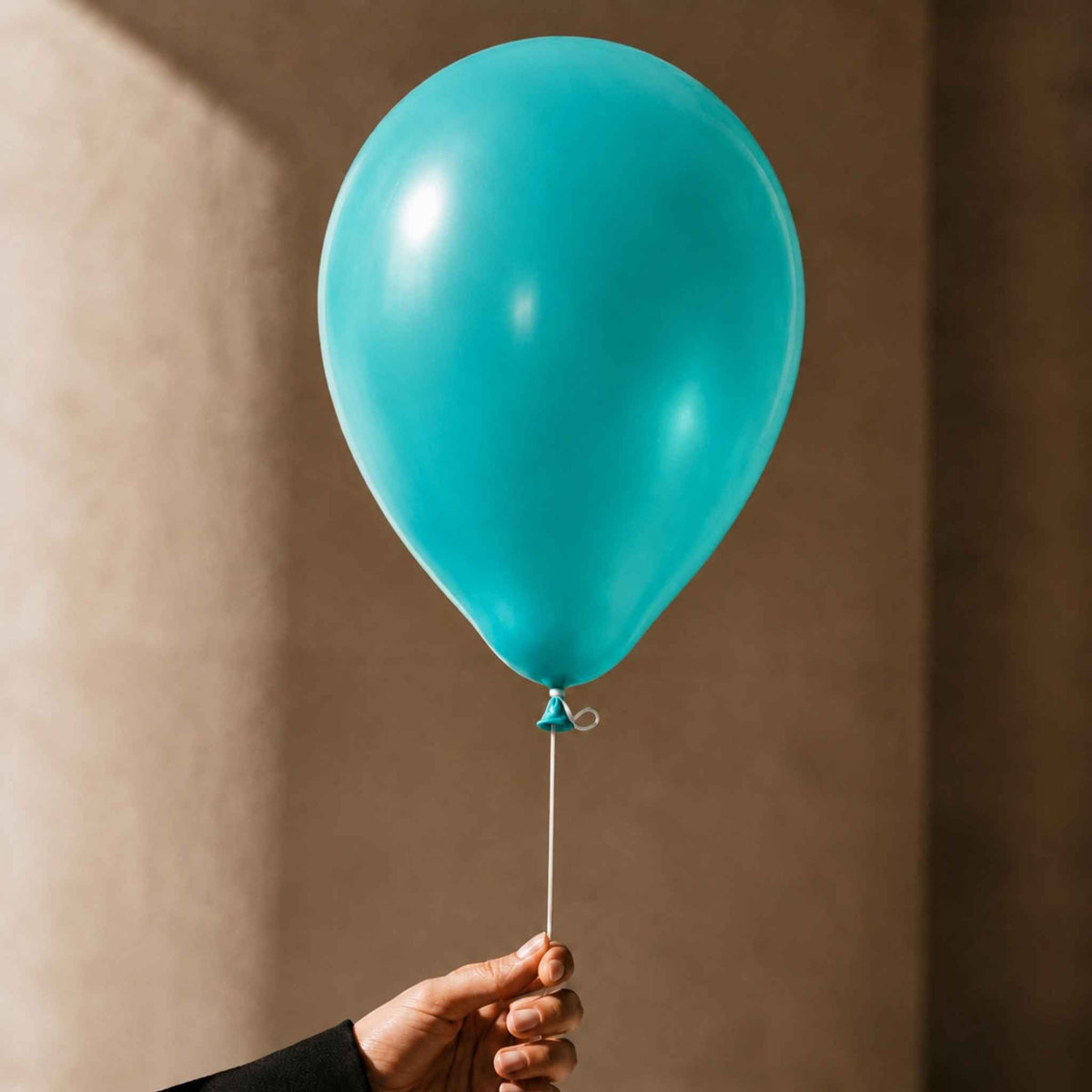 A single inflated teal balloon held by a person's hand against a warm beige wall.