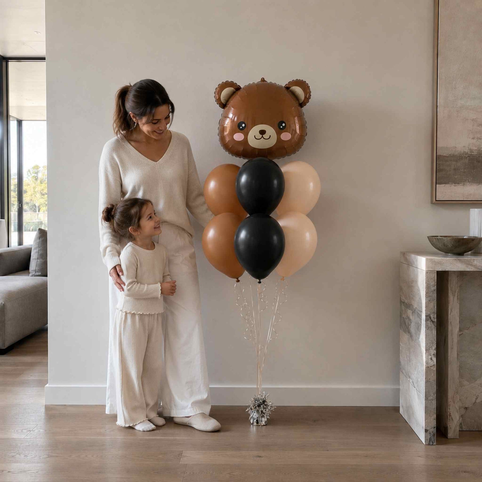 Woman and child standing next to a large teddy bear-shaped balloon in a living room.