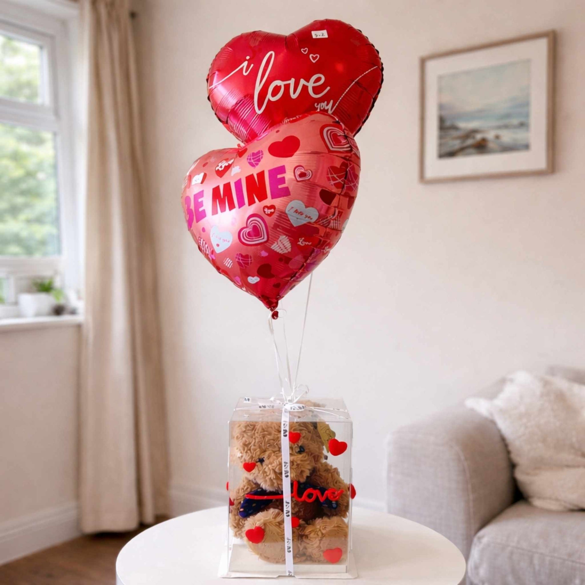 Heart-shaped balloons with 'I love you' and 'SHE'S MINE' text, teddy bear in a box, on a table in a room.