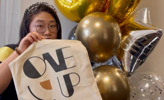 A person holding up a cream canvas tote bag printed with the words 'ONE UP' in bold black letters, with a small orange accent shape. The person is wearing a black 'BIRTHDAY' headband, and the background features a cluster of gold and silver balloons.