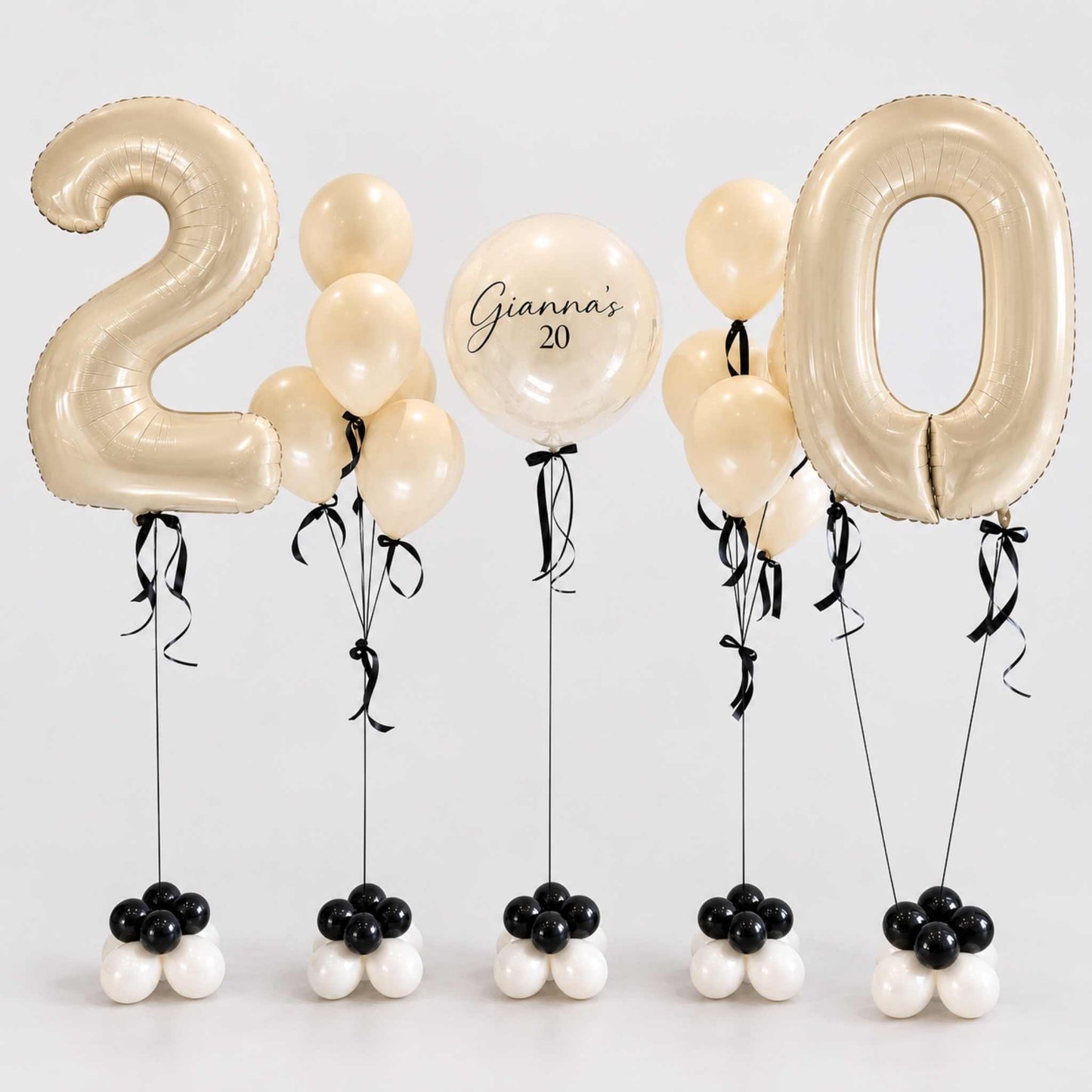 A set of birthday balloons spelling out '20' for a 20th birthday celebration. The set includes two large metallic gold foil balloons shaped like the numbers '2' and '0', several cream-colored latex balloons, one round balloon with the text 'Gianna's 20', and five small balloon clusters at the bottom in black, white, and cream.