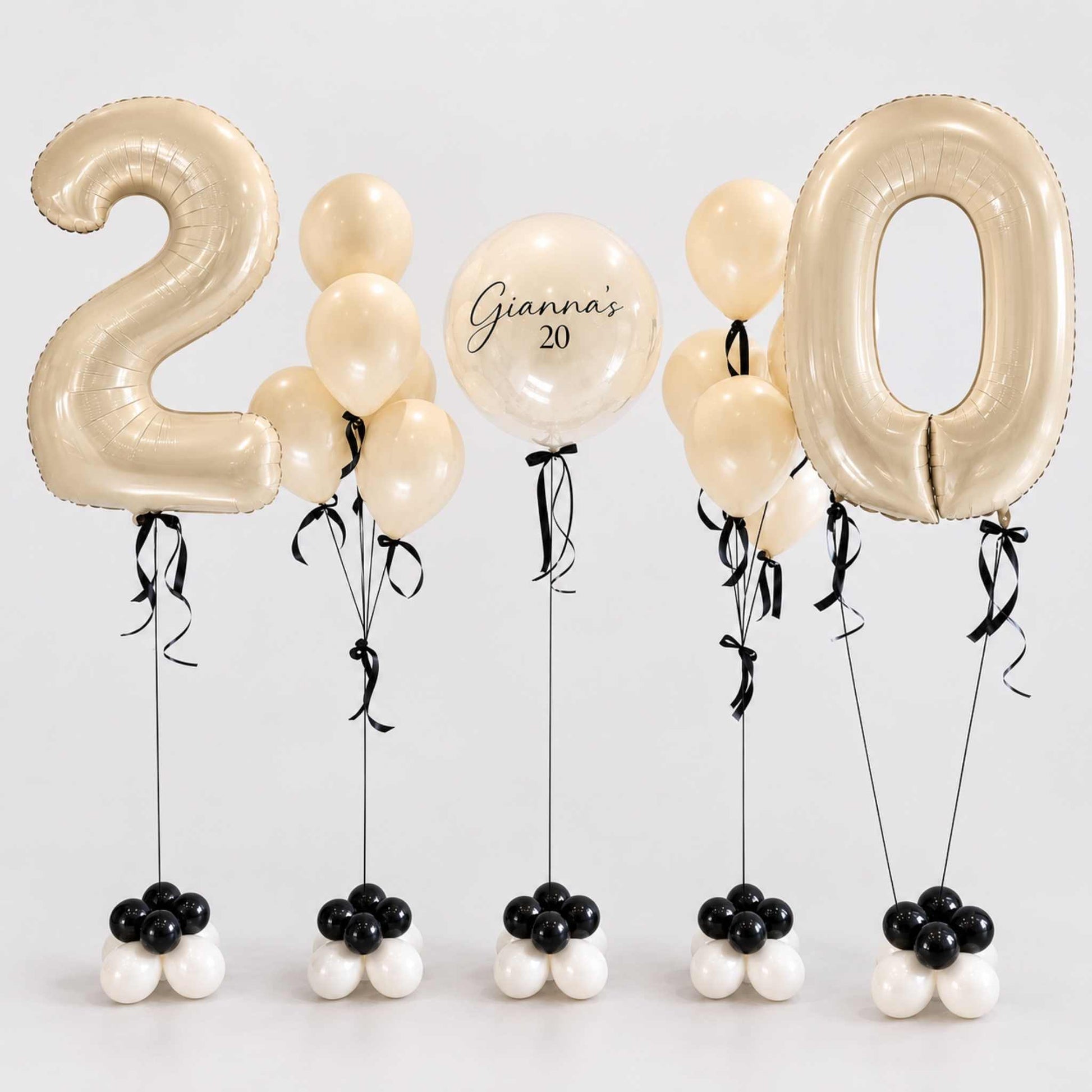 A set of birthday balloons spelling out '20' for a 20th birthday celebration. The set includes two large metallic gold foil balloons shaped like the numbers '2' and '0', several cream-colored latex balloons, one round balloon with the text 'Gianna's 20', and five small balloon clusters at the bottom in black, white, and cream.