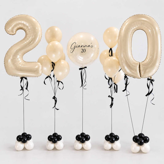 A set of birthday balloons spelling out '20' for a 20th birthday celebration. The set includes two large metallic gold foil balloons shaped like the numbers '2' and '0', several cream-colored latex balloons, one round balloon with the text 'Gianna's 20', and five small balloon clusters at the bottom in black, white, and cream.