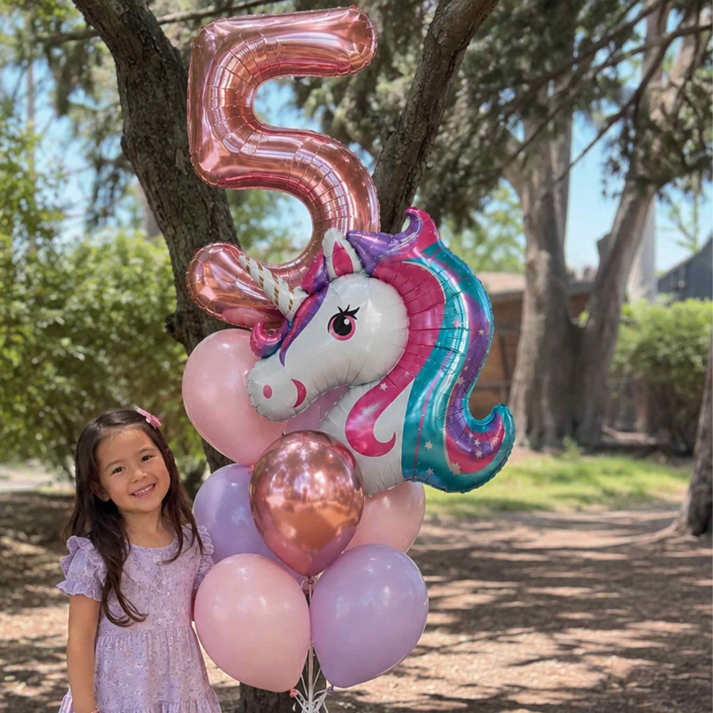 A young girl in a lavender dress stands outdoors next to a cluster of foil balloons, including a large metallic rose-gold number 5 balloon, a unicorn head balloon with pink, purple, and teal coloring, and several pastel pink and lavender balloons. Trees and a sunny backyard setting are visible in the background.