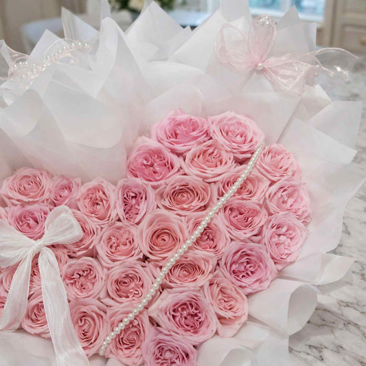 Heart-shaped arrangement of pink roses with white ribbons and pearl accents on a marble surface.