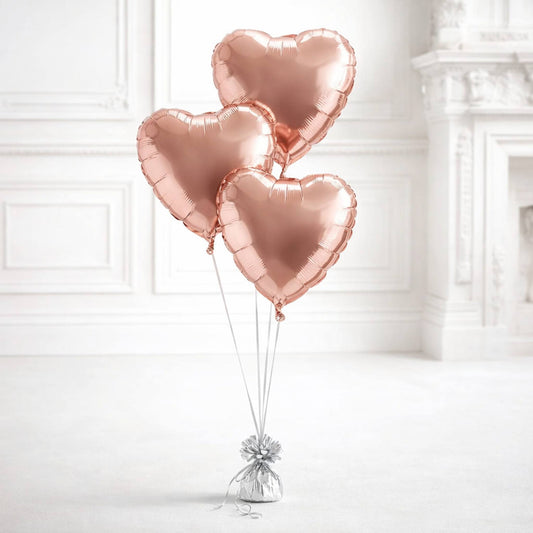 Three rose gold heart-shaped balloons tied together against a white wall.