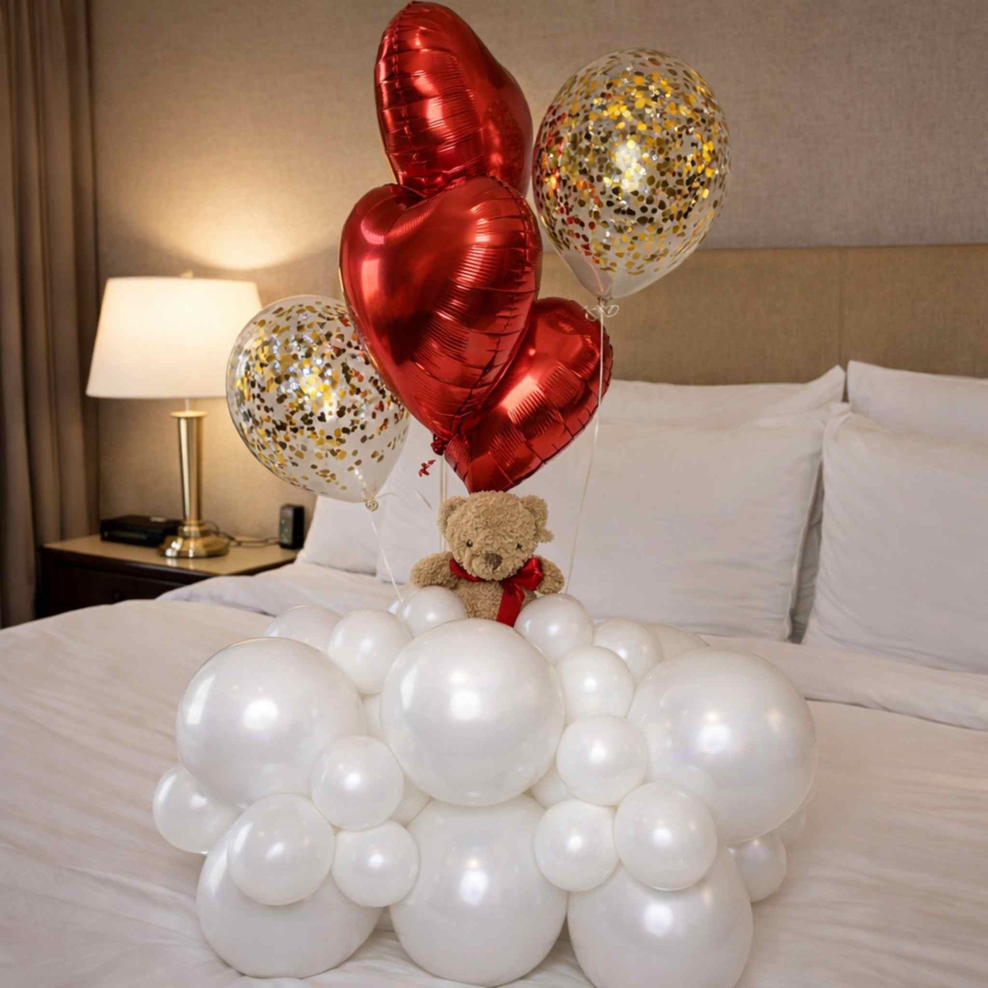 Decorative setup with red heart-shaped balloons, white balloons, and a teddy bear on a bed.